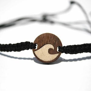 May include: A close-up of a bracelet featuring a round, dark wood charm with a wave design. The wave is light-colored and set against the dark wood. The bracelet has a black, braided cord and a white cord. The bracelet is on a white background.