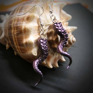 May include: A pair of dangling earrings featuring purple octopus tentacles with visible suction cups. The tentacles are attached to silver-toned metal fishhook ear wires, creating a curved, organic shape that hangs downward from the top.