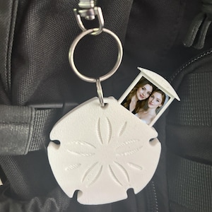 May include: A white sand dollar-shaped keyring with a silver ring, attached to a black backpack. The sand dollar has a detailed design and a small compartment that opens to reveal a photo of two women.