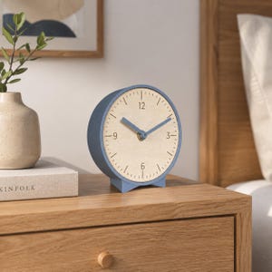 May include: A blue and beige analog clock with a round face and a light blue frame. The clock has a beige face with black numbers and blue hands. The clock is sitting on a wooden nightstand.