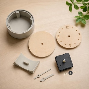 May include: Clock-making kit with components laid out on a light wood surface. Includes a gray plastic casing, wooden clock faces, clock hands, and a black clock mechanism. The clock face has the numbers 3, 6, 9, and 12.