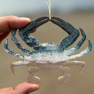 May include: A teal blue and white crab ornament with a string for hanging. The crab is painted to look like a wave crashing on the beach.