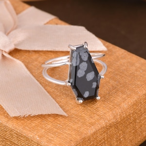 Puede incluir: Un anillo de plata con una piedra preciosa de obsidiana copo de nieve en forma de ataúd, con una base negra e inclusiones circulares grises. La piedra está sujeta por cuatro garras en una banda de plata fina y pulida que se divide en dos.