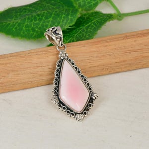 Op de afbeelding: Een druppelvormige hanger met een gladde, lichtroze steen, gezet in een gedetailleerd montuur van geoxideerd sterling zilver met krulwerk en kraalversieringen, inclusief een decoratief zilveren oogje aan de bovenzijde voor bevestiging.