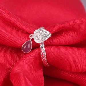 May include: A silver ring featuring a textured band and a heart-shaped top element. A small, teardrop-shaped pink gemstone charm hangs from the side of the heart. The metal surface of the ring and the heart component has a hammered, reflective finish.