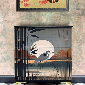 May include: A black chest of drawers with gold accents and a painted scene of a heron in a marsh with a full moon. The chest of drawers has four drawers and sits on gold legs.
