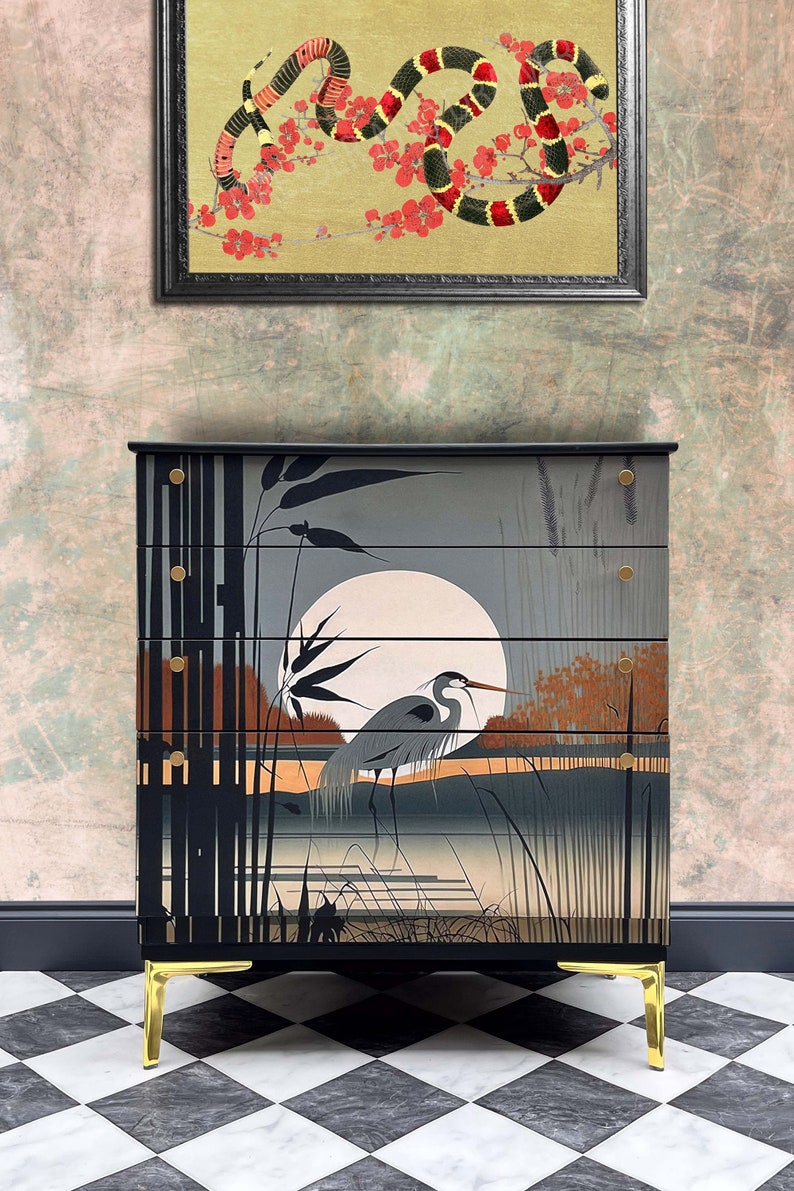 May include: A black chest of drawers with gold accents and a painted scene of a heron in a marsh with a full moon. The chest of drawers has four drawers and sits on gold legs.