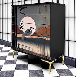 May include: A black chest of drawers with four drawers featuring a hand-painted design of a heron standing in a marsh with a full moon in the background. The chest has gold-colored legs.