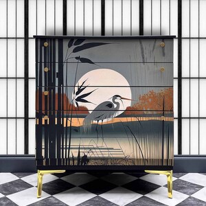 May include: A black chest of drawers with four drawers features a hand-painted design of a heron standing in a marsh under a full moon. The chest has gold-colored legs.