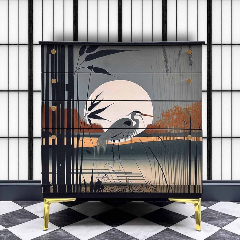May include: A black chest of drawers with four drawers features a hand-painted design of a heron standing in a marsh under a full moon. The chest has gold-colored legs.