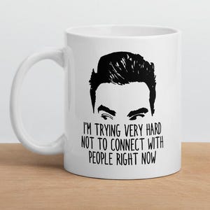 May include: White ceramic mug with a black graphic of a person's face and the text "I'M TRYING VERY HARD NOT TO CONNECT WITH PEOPLE RIGHT NOW". The mug is on a wooden surface.
