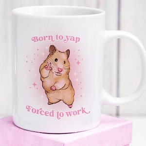 May include: White ceramic mug with a cartoon hamster illustration. The hamster is light brown with pink paws and ears, giving a peace sign. The mug has the text "Born to yap" and "Forced to work" in pink script.