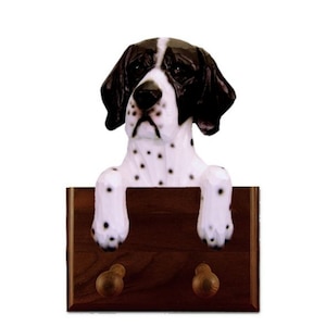 May include: A black and white English Pointer dog figurine with brown eyes, peeking over a wooden coat rack with two hooks. The figurine is made of painted resin and has a realistic appearance.