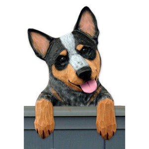 May include: A wooden figurine of a blue heeler dog peeking over a fence. The dog has black, blue, and brown markings and is smiling with its tongue sticking out.