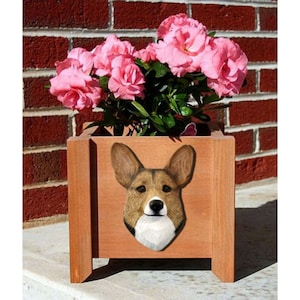 May include: A wooden planter box with a brown and white corgi dog face painted on the front. The planter is filled with pink flowers.