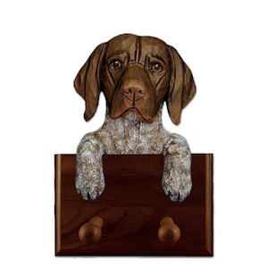 May include: A wooden coat rack featuring a German Shorthaired Pointer dog head with brown and white fur. The dog is looking forward with its paws resting on the top of the rack. The rack has two wooden hooks.