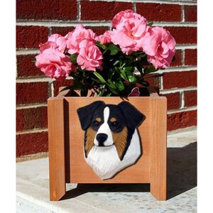 May include: A wooden planter box with a brown, black, and white dog head sculpture on the front. The planter is filled with pink flowers and green leaves.