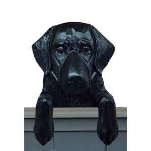 May include: Black Labrador Retriever dog figurine peeking over a ledge. The dog is carved from wood and has a realistic appearance.