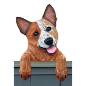 May include: A wooden figurine of a red and white Australian Cattle Dog peeking over a fence. The dog has a pink tongue sticking out and is looking at the camera.