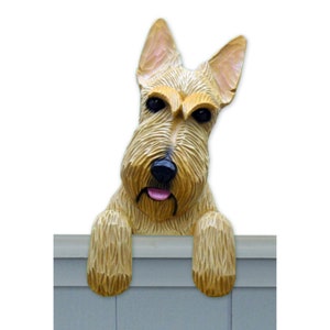 May include: A wooden figurine of a Scottish Terrier dog peeking over a fence. The dog is light brown with black eyes and a pink tongue. The figurine is hand-painted and has a realistic appearance.