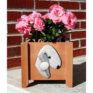 May include: A wooden planter box with a carved Bedlington Terrier dog head on the front. The planter is filled with pink flowers and is sitting on a brick surface.