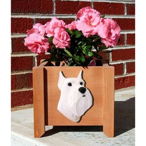 May include: A white wooden planter box with a white Schnauzer dog head carved into the front. The planter is filled with pink flowers.