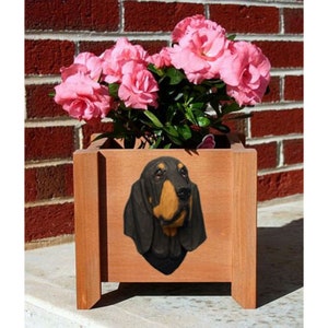 May include: A wooden planter box with a black and tan Bloodhound dog head sculpture on the front. The planter is filled with pink flowers.
