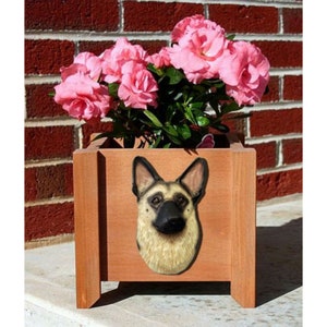 May include: A wooden planter box with a brown finish features a sculpted German Shepherd dog head on the front. The planter is filled with pink flowers.