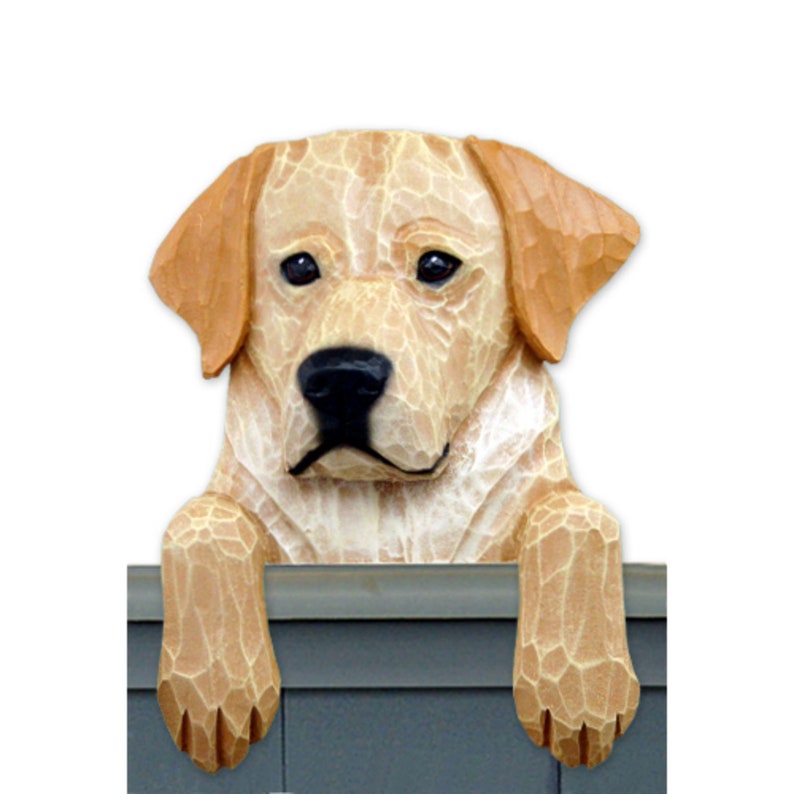 May include: A wooden figurine of a yellow Labrador Retriever peeking over a ledge. The dog has a light brown coat and is looking at the viewer with its head tilted slightly to the side.