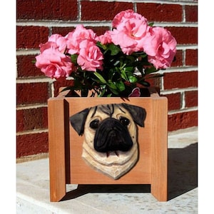 May include: Wooden planter box with a carved pug dog face on the front. The planter is filled with vibrant pink azalea flowers and green foliage. The box is made of light brown wood and has a rectangular shape.