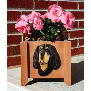May include: A wooden planter box with a black and tan dog head carved into the front. The planter is filled with pink flowers and green leaves.