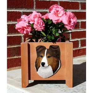 May include: A wooden planter box with a carved image of a brown and white Shetland Sheepdog on the front. The planter is filled with pink flowers and is sitting on a brick surface.