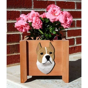 May include: A wooden planter box with a brown and white dog head sculpture on the front. The dog has a white muzzle and a brown and white coat. The planter is filled with pink flowers.