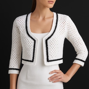 May include: A white crocheted cardigan with black trim around the neckline, hem, and sleeves. The cardigan is cropped and has a V-neck.