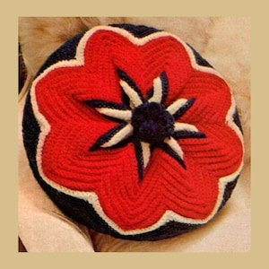 Puede incluir: Un cojín redondo hecho a mano con ganchillo con un diseño floral. El cojín presenta una forma de flor roja con detalles en blanco y azul marino. El centro de la flor tiene un pompón azul marino. El cojín mide aproximadamente 35 cm de diámetro.