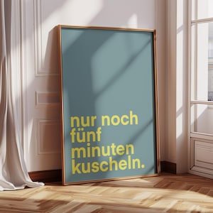 Könnte beinhalten: Gerahmtes Poster mit türkisblauem Hintergrund und goldenem Text, der lautet: "nur noch fünf minuten kuscheln." Das Poster befindet sich in einem goldenen Rahmen und lehnt an einer weißen Wand mit Holzboden.