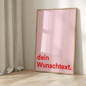 Puede incluir: Una impresión artística rosa enmarcada con el texto alemán "dein Wunschtext." en rojo. La obra de arte tiene un acabado brillante y se exhibe contra una pared blanca y un suelo de madera. El marco es de color madera clara.