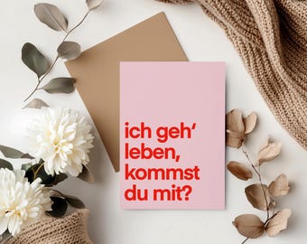 May include: A pink card with the text "ich geh' leben, kommst du mit?" in red letters. The card is on a white surface with brown branches and a brown knitted blanket.