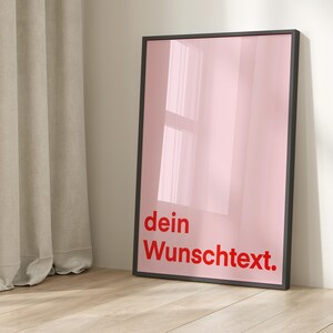 May include: A framed pink poster with the words "dein Wunschtext" in red. The poster has a black frame and is leaning against a white wall. The floor is light-colored wood.