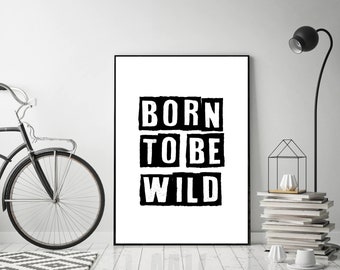 May include: Black framed art print with the words "BORN TO BE WILD" in bold, block letters. The artwork is displayed against a white wall, with a black bicycle and a modern black lamp in the background. A stack of books and decorative objects are also visible.