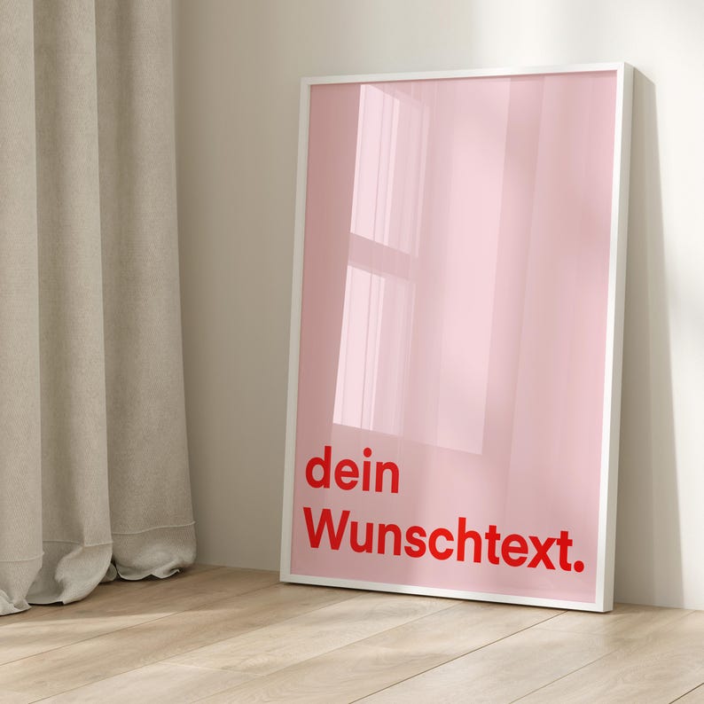 May include: A framed pink art print with the words "dein Wunschtext." in red. The artwork has a glossy finish and is displayed in a white frame. The print is leaning against a white wall.