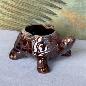 Może przedstawiać: Ceramiczna popielniczka w kształcie żółwia w odcieniach brązu z błyszczącym wykończeniem. Popielniczka ma głęboki, okrągły otwór i jest podparta przez cztery małe nóżki. Skorupa żółwia ma teksturowane detale.