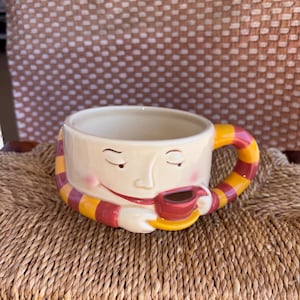 May include: A whimsical ceramic coffee mug with a smiling face design. The mug features a cream-colored base, a red and yellow striped handle, and a matching scarf design. The mug is holding a small red cup of coffee.