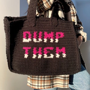 Könnte beinhalten: Schwarze Häkeltasche mit den Worten "DUMP THEM" in Pink und Weiß. Die Tasche hat zwei Henkel und ist aus dickem, strukturiertem Garn gefertigt. Die Tragetasche ist ein handgefertigter Artikel.
