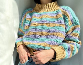 May include: A striped crocheted sweater with a high neck in shades of beige, purple, and teal. The sweater is worn over blue jeans.