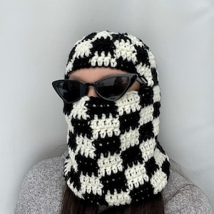 Könnte beinhalten: Eine schwarz-weiße karierte Häkelmütze mit einem schwarzen Häkelrand. Die Mütze wird von einer Person mit einer schwarzen Sonnenbrille getragen.