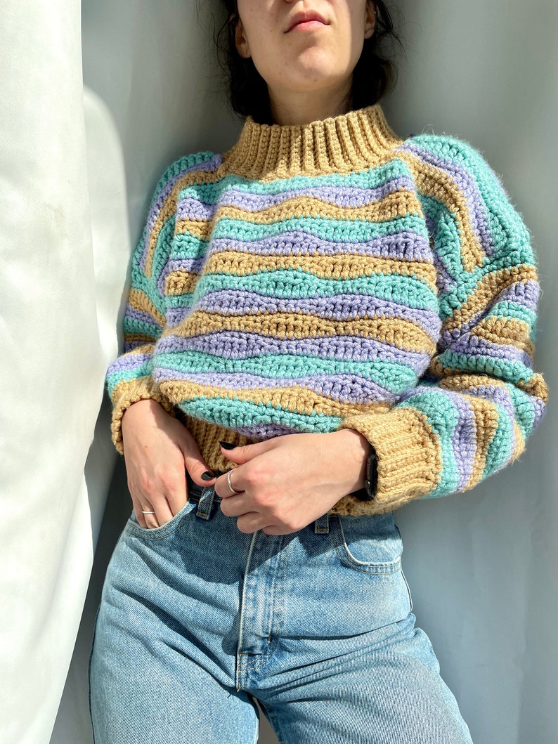Op de afbeelding: Een gestreepte gehaakte trui met een hoge hals in de kleuren beige, paars en turquoise. De trui wordt gedragen over een blauwe jeans.