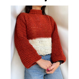 Op de afbeelding: Een roestkleurige gebreide trui met een witte gebreide band aan de onderkant. De trui heeft een hoge hals en lange mouwen.
