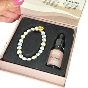 May include: A gift set featuring a white and gray beaded bracelet with gold accents and a small bottle of patchouli essential oil. The set is presented in a pink box with text that reads "LAVA BEADS" and "Wallace & Green Patchouli".