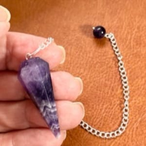 May include: A purple amethyst crystal pendulum with a silver chain and a small amethyst bead at the end.